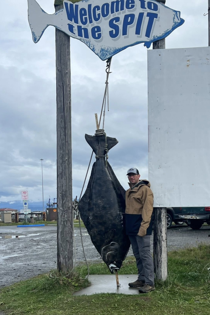 Welcome to the Spit Halibut Photo – Homer, Alaska - Dohrn Travels Man standing beside a large halibut at the “Welcome to the Spit” sign in Homer, Alaska.