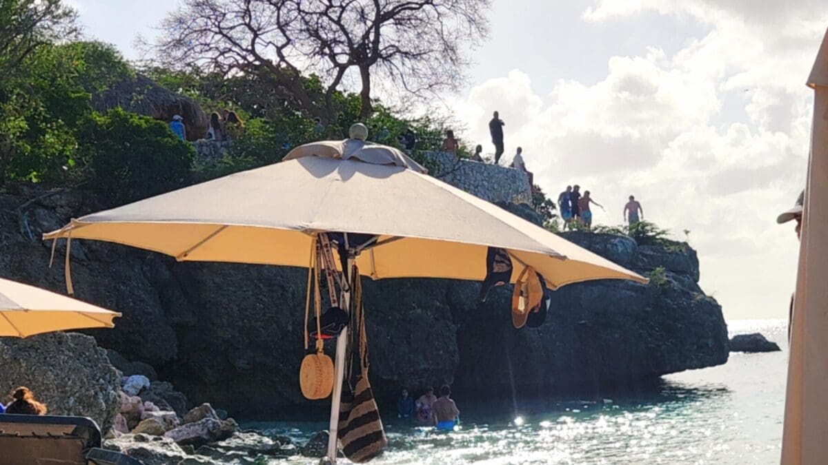 Cliff Jumping at Grote Knip Beach Curaçao - Dohrn Travels People cliff jumping into the water at Grote Knip Beach in Curaçao with umbrellas and rocky shoreline